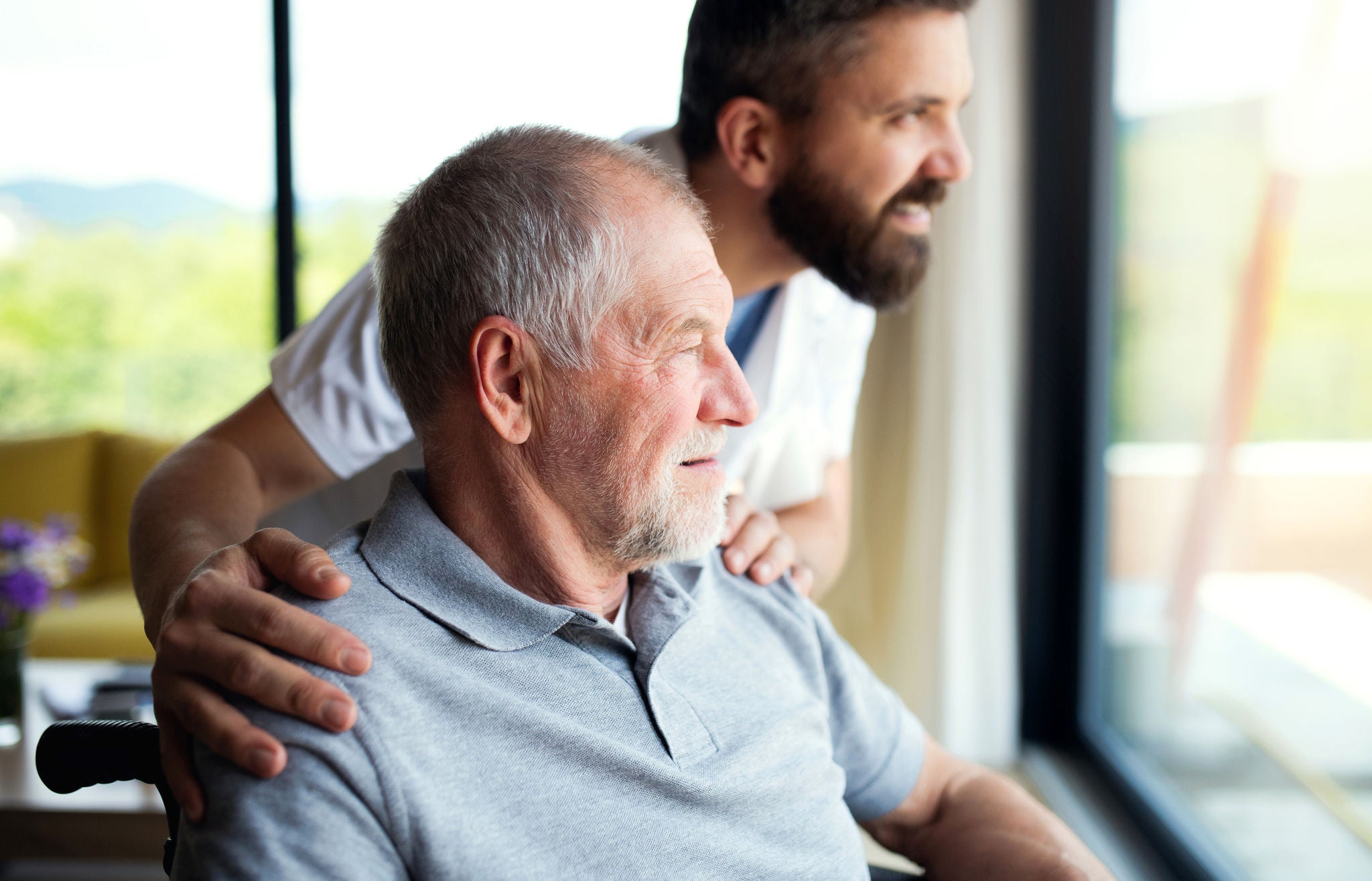 A mature man caregiver with stethoscope and old patient looking out through window.