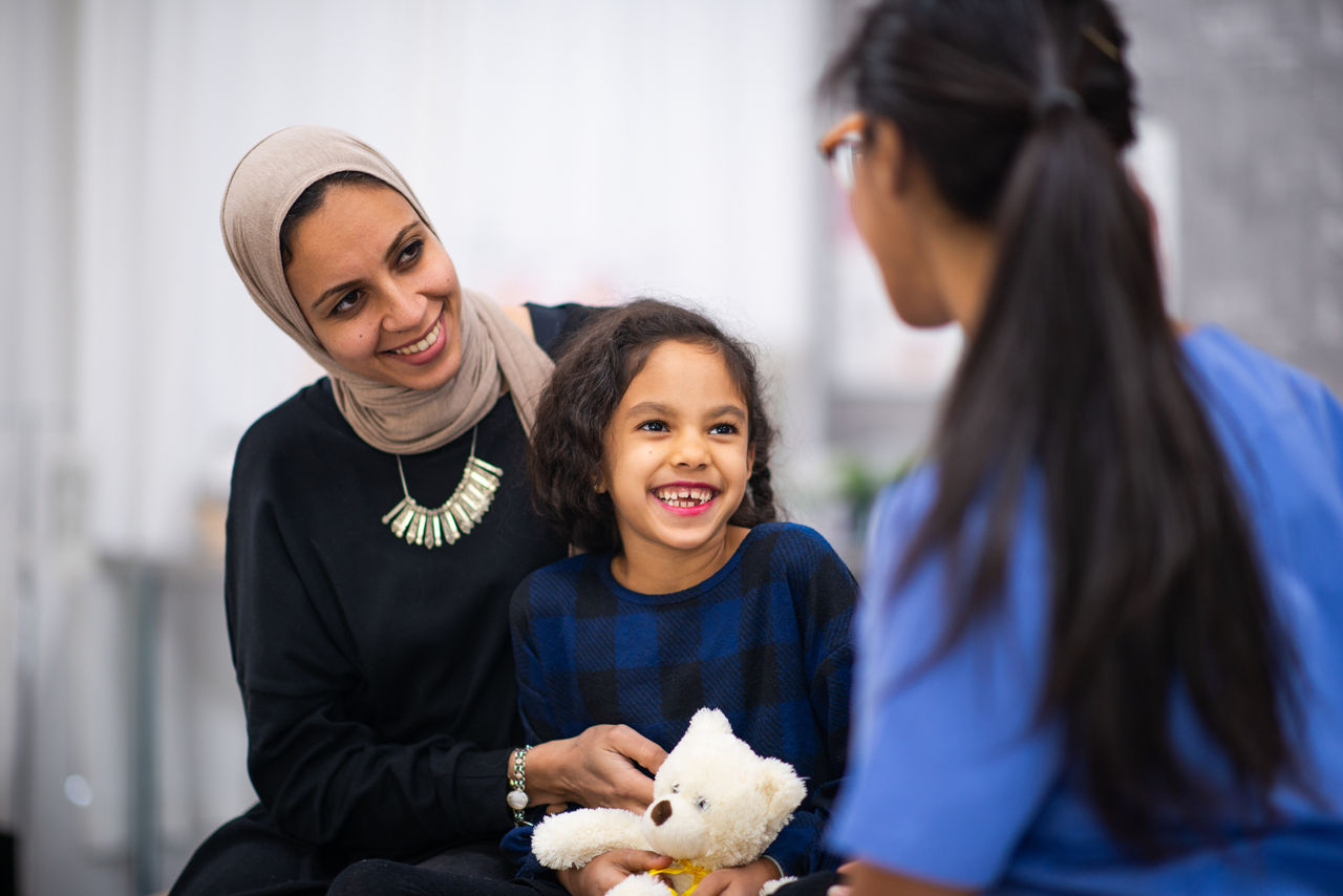 A little girl sits on her Mothers lap on the examination table, while on a visit to the doctor. Her mother is wearing a head scarf and both are casually dressed. The little girl is holding a white teddy bear with a yellow ribbon and looking up at her East Indian female doctor. The doctor has her back to the camera as the mother explains her daughters symptoms.